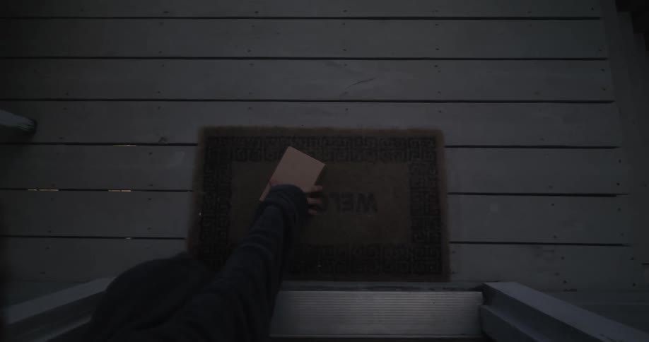 A person reaches out of their doorway and picks a package up off the mat on the porch