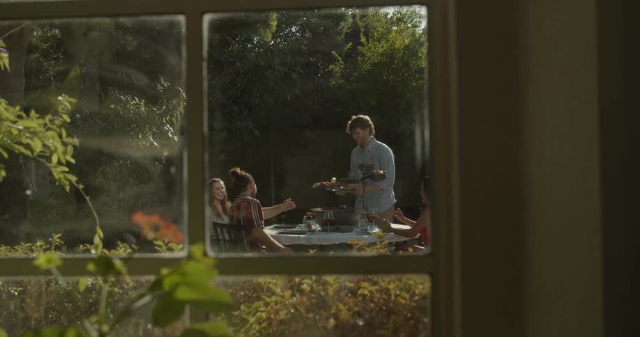 Group of friends eating on a table in the backyard at morning seen through a window