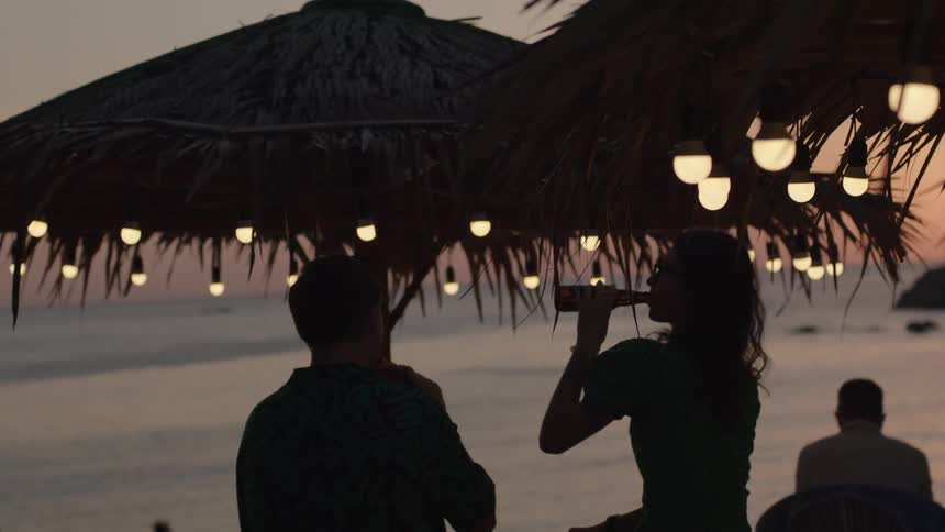 A man and woman at a bar on the beach drink beer together