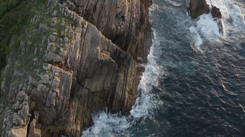 Ocean waves splash against the rocky cliffs as hikers climb a ridge on the Spanish Atlantic shore