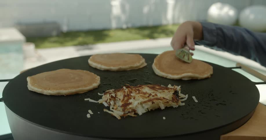 A person cooking pancakes and hash brown over a pan in the backyard near the pool