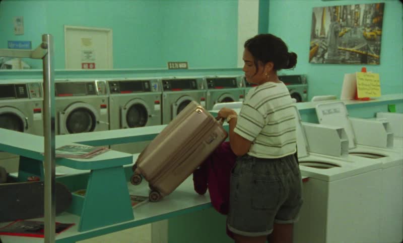 Young woman carrying luggage enters a laundromat and starts unpacking clothes while talking to a person