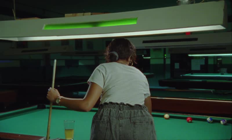 A woman is standing next to a pool table holding her cue stick as she sets her beer down to rack the balls