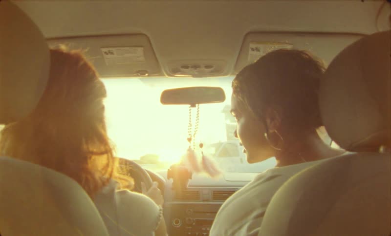 The sun shines brightly upon an automobile as two women sitting inside are talking as they travel on the road