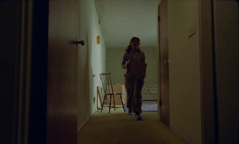 A woman is walking out of a room and down a hallway carrying a box
