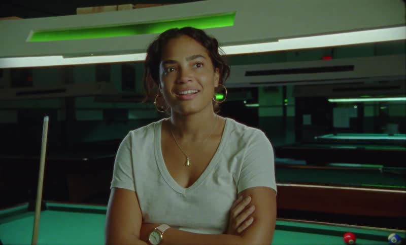 Young woman reclined on a billiard table listening to someone talking out of frame while laughing