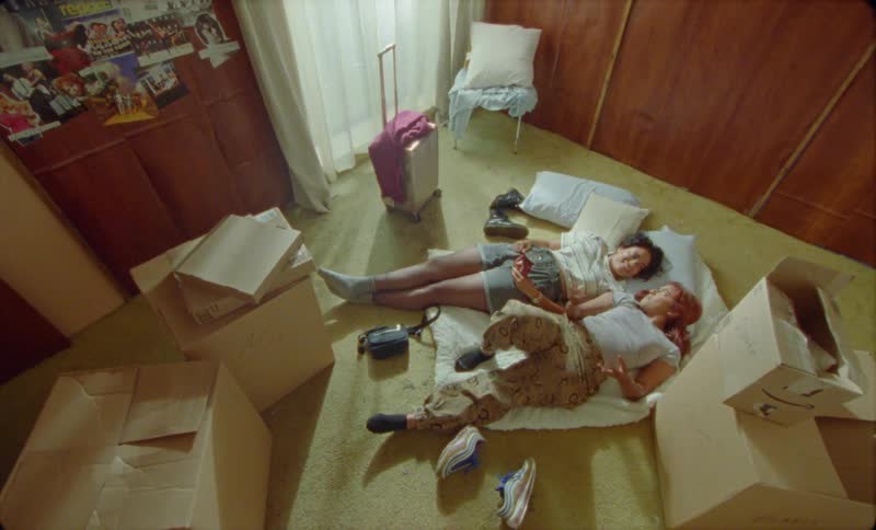 Two happy friends talk while resting on a futon on the floor of a bedroom surrounded by cardboard boxes