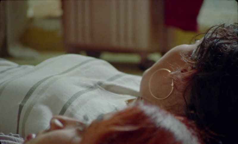 Two women lie in bed together and one turns and looks at a suitcase on the floor