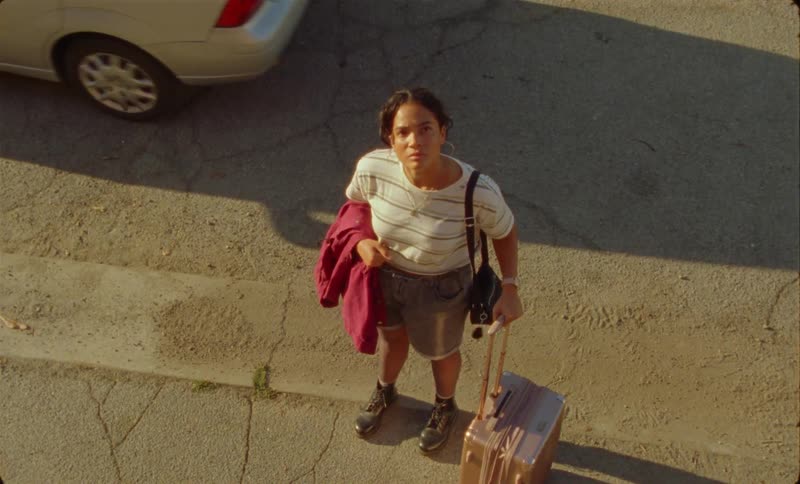 Car pulls away as woman rolls suitcase forward before stopping to stare upward and smiling
