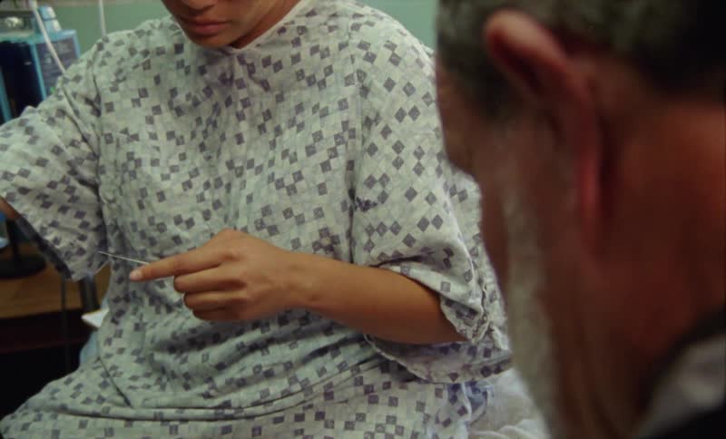 Young tired woman wearing a hospital gown sitting on a bed listening to a doctor