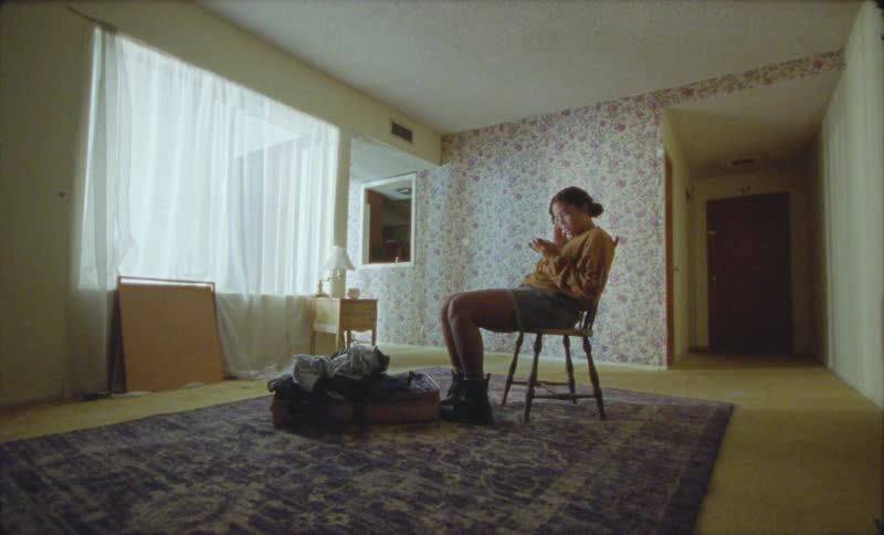 Woman with bun yellow sweater shorts and boots sits on chair answering phone call in front of open suitcase