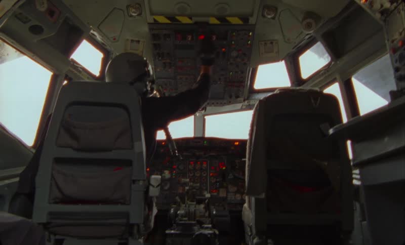 A pilot in the cockpit looks panicked as he pushes buttons and controls