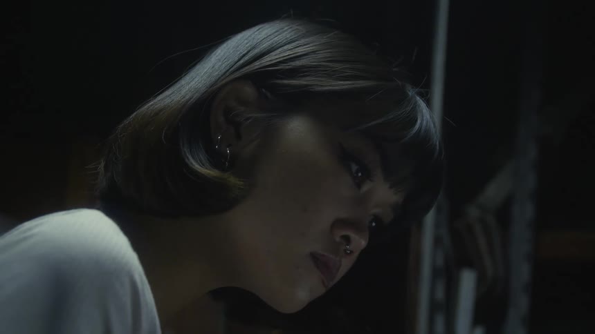 Woman with bob hairstyle earrings piercing white shirt and tattoos works on laptop in dark room