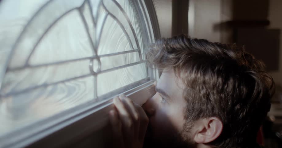 There is a young white man that is looking outside through the window