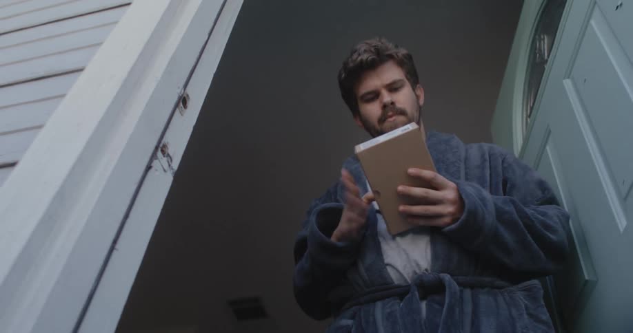 Man with beard and bathrobe picks up small package from doormat