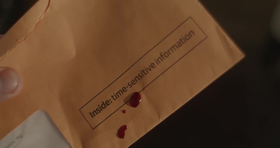 Blood drops fall on to a manilla envelope as someone holds it in their hand