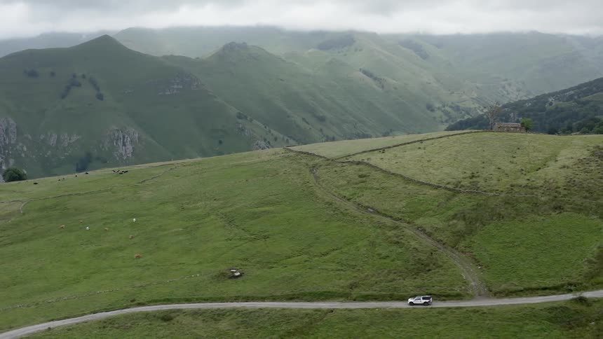 A truck travels on a country road near farm animals grazing and mountains in the distance under a hazy sky in the north of Spain