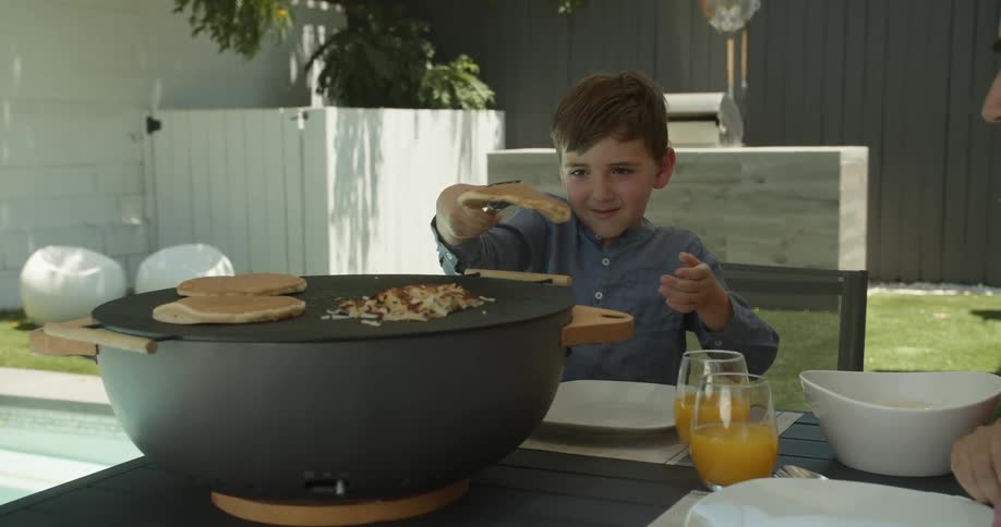A boy serves himself a pancake from the tabletop griddle while sitting on the patio with dad