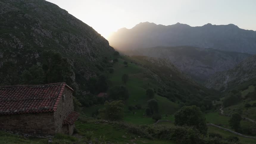 The sun shines behind a green mountain slope over a stone cabin