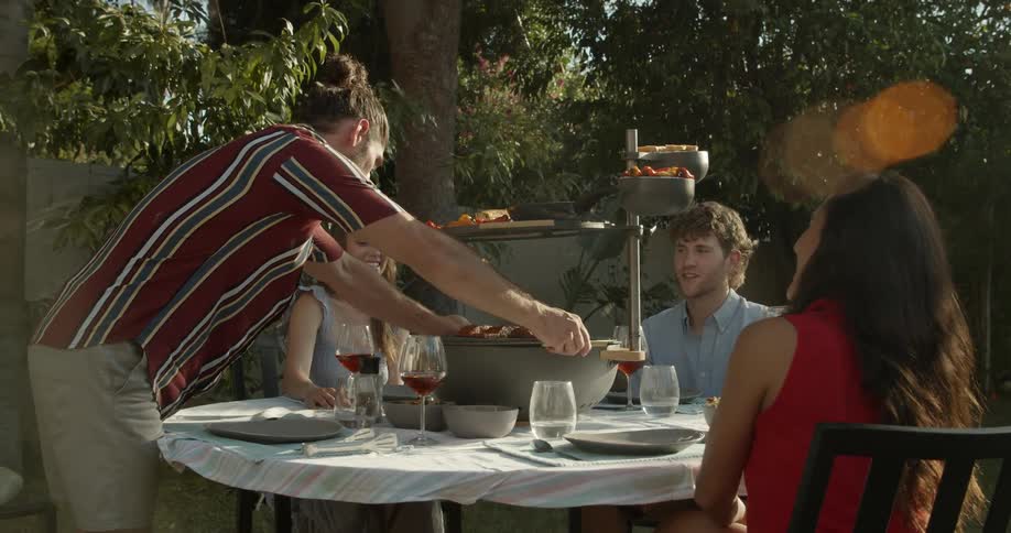 Man places food onto multilayered serving holder in middle of outdoor table as friends smile