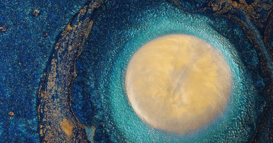 An organic blue and gold substance sparkles as it moves and a drop falls creating a vortex that starts with an orb and a sphere emerges and goes back down as it is covered