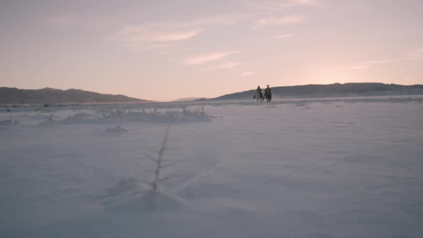 Two riders on horseback cross a snowy field