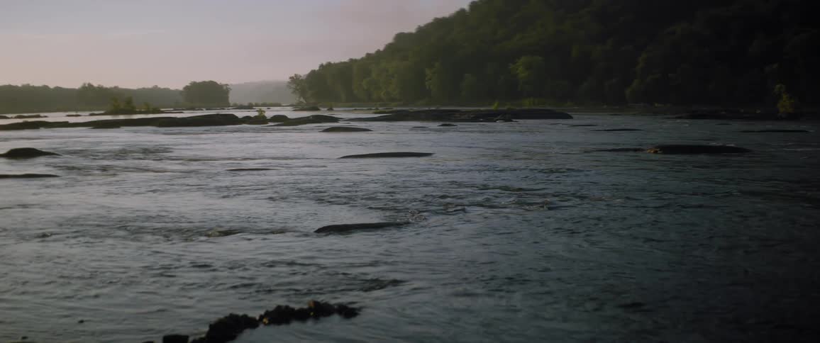 River water flows over rocks and a forest is along the coast with a mountain in the distance under a hazy sky