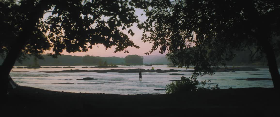 A man is seen through the trees standing in the water close to shore fishing