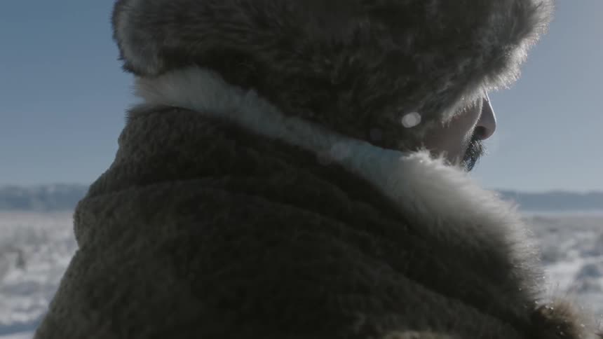 A man in heavy clothing and a fur hat stands in the snow on a cold sunny day