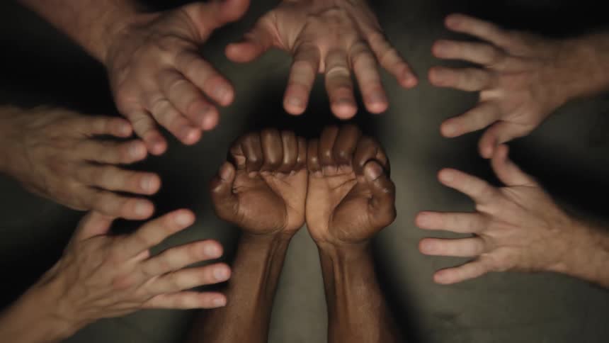A black person has their arms out and closes their fingers into a fist and several white people move their open hands to the black fists