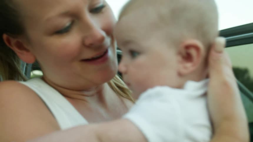 Blonde mother with ponytail takes baby from stroller and kisses baby next to gray car
