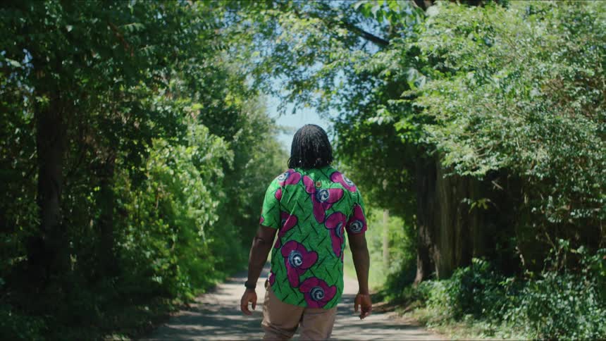 Man with dreadlocks green t shirt brown pants walks along road in the forest