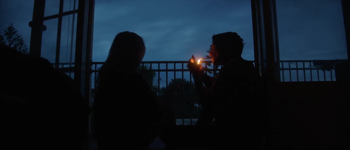 Two people are sitting outside on a balcony and the woman lights her cigarette and she passes her lighter to the man and he lights his cigarette