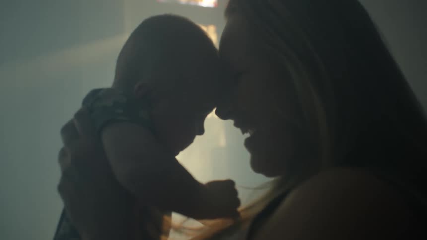 Mother smiles as she holds baby in dim room lit by rays from the window
