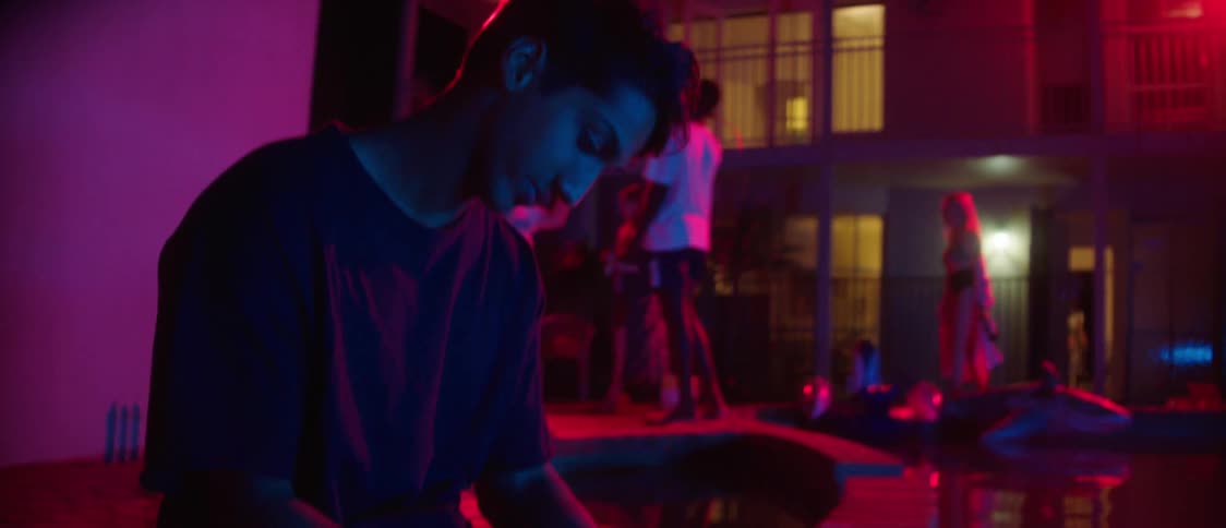 A man is sitting on the edge of a pool with a beer bottle as people party behind him