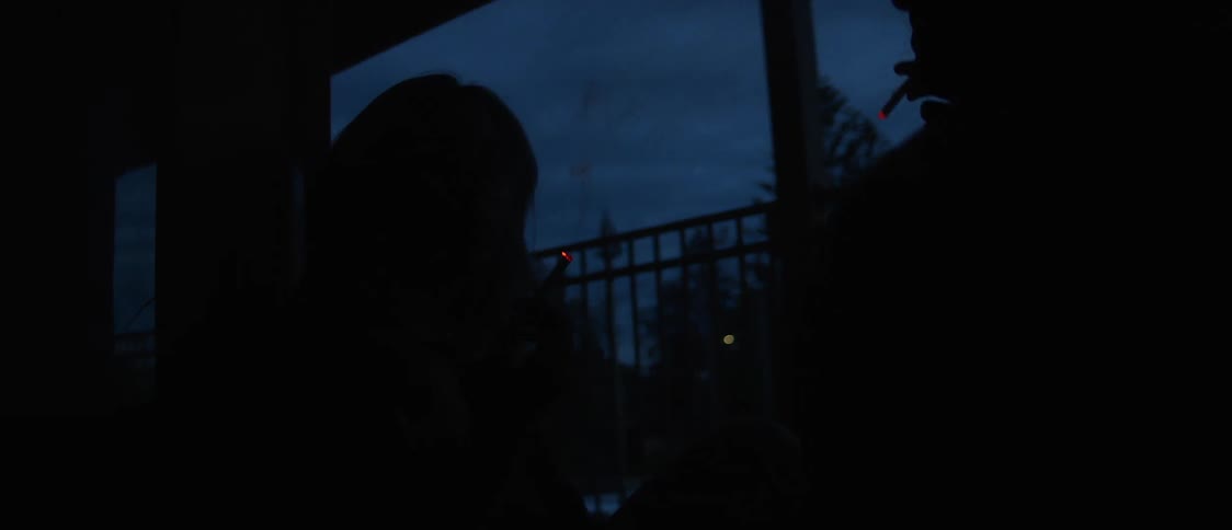 A man lights a cigarette for a woman as they stand on a porch