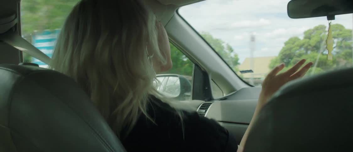 Blonde woman with black t shirt dances and talks to man driving car in the street