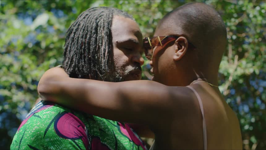 Man with dreadlocks beard and green shirt makes out woman with shaven head and sunglasses in the forest