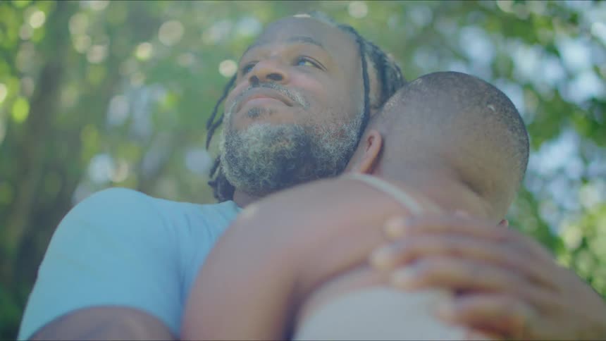 A man and woman are standing outside near trees and her head is leaning on his shoulder as his arms are around her