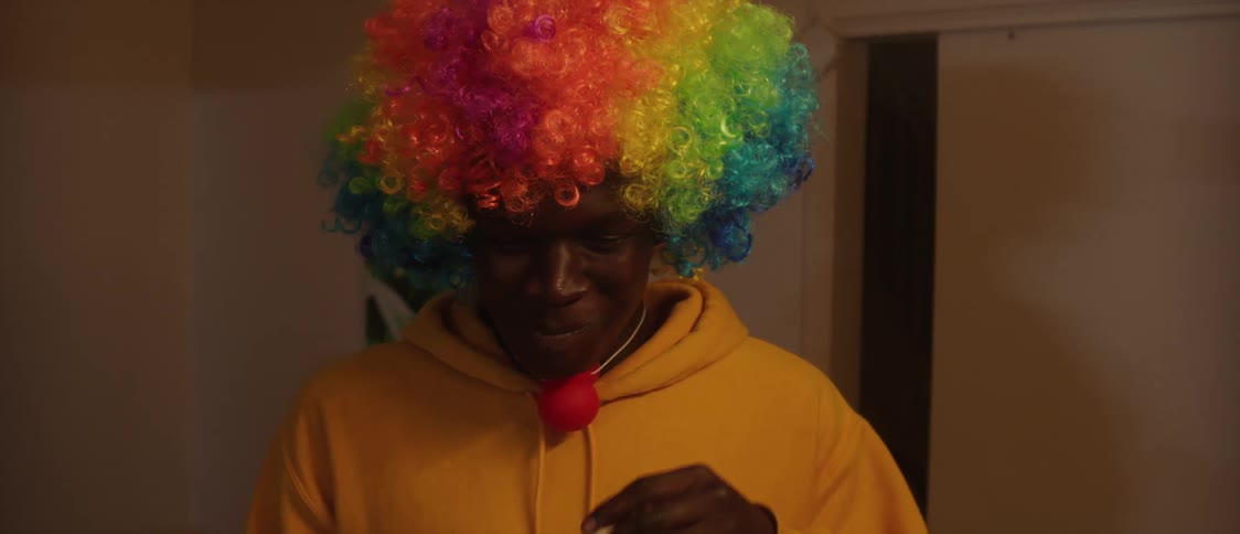 Man with curly wig with rainbow and yellow hoodie smokes cigarette laughing