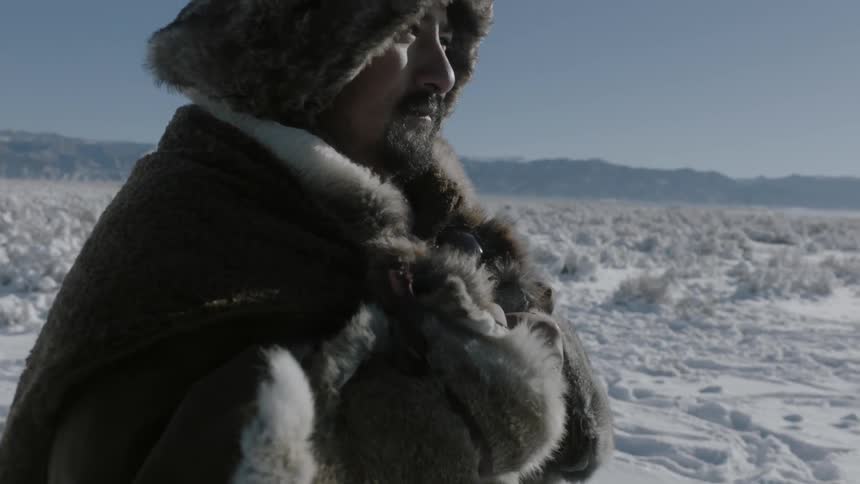 A man wearing heaving clothing and a fur hat stands outside in the snow
