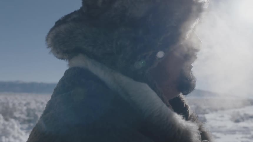 A man in heavy clothing and a fur hat stands in the snow on a cold sunny day