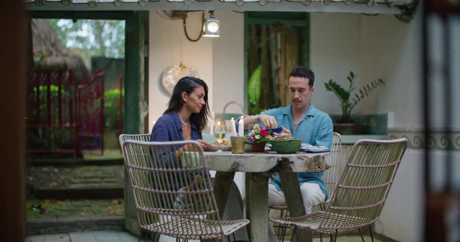Man seasons meals as he and woman sit at table on patio