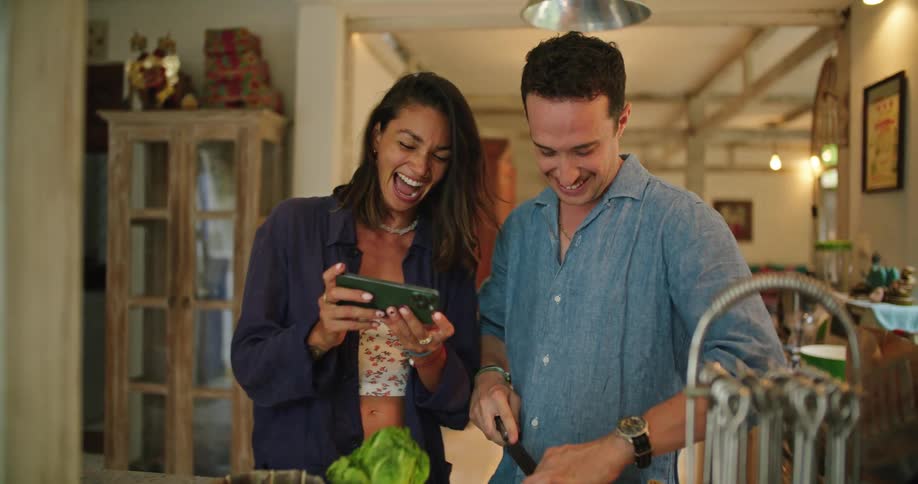 Young happy couple cooking at home laugh while looking at pictures on a phone