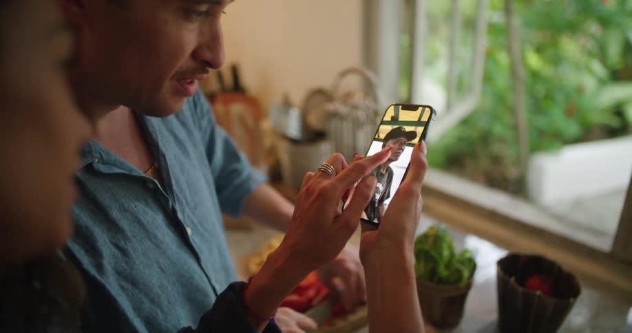 Man chopping vegetables in the kitchen looks at pictures his partner shows him in the phone