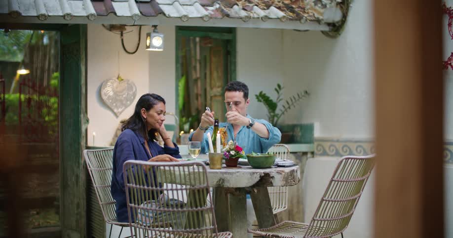 A man and woman are having a romantic candlelit dinner outside of their home and the man serves the woman spaghetti