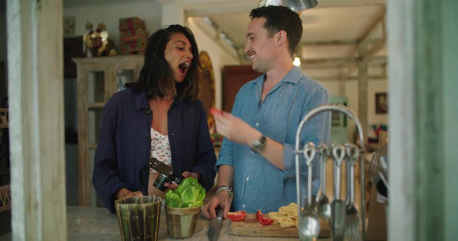 Man chopping vegetables at home puts a slice of tomato on his wife mouth