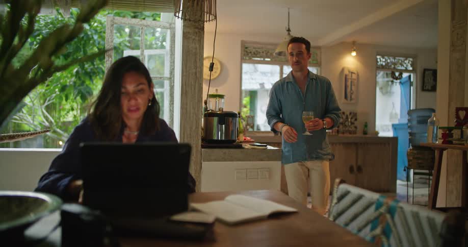 Man serves a glass of wine to his wife browsing a laptop in the dinning room
