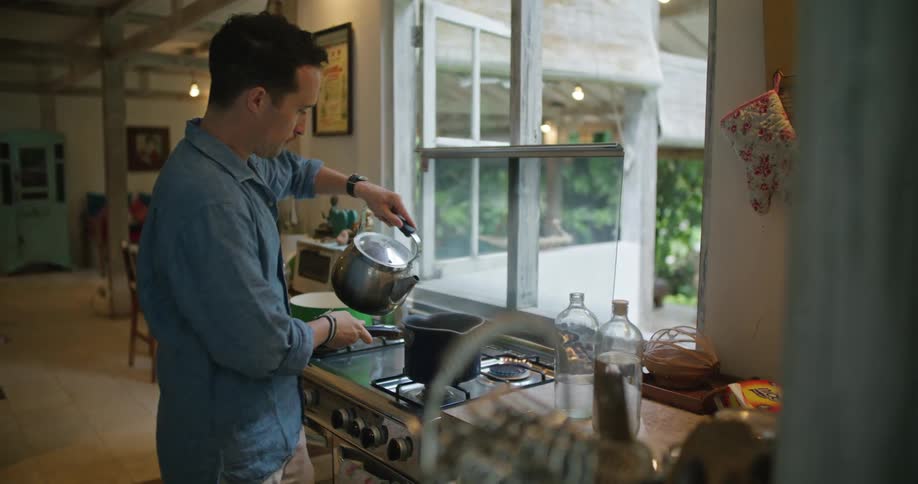 A man is standing over a stove pouring water from a kettle into a pot