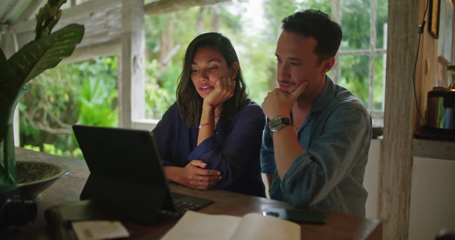 A man and a woman are sitting together at a table with a laptop computer talking and looking at the screen